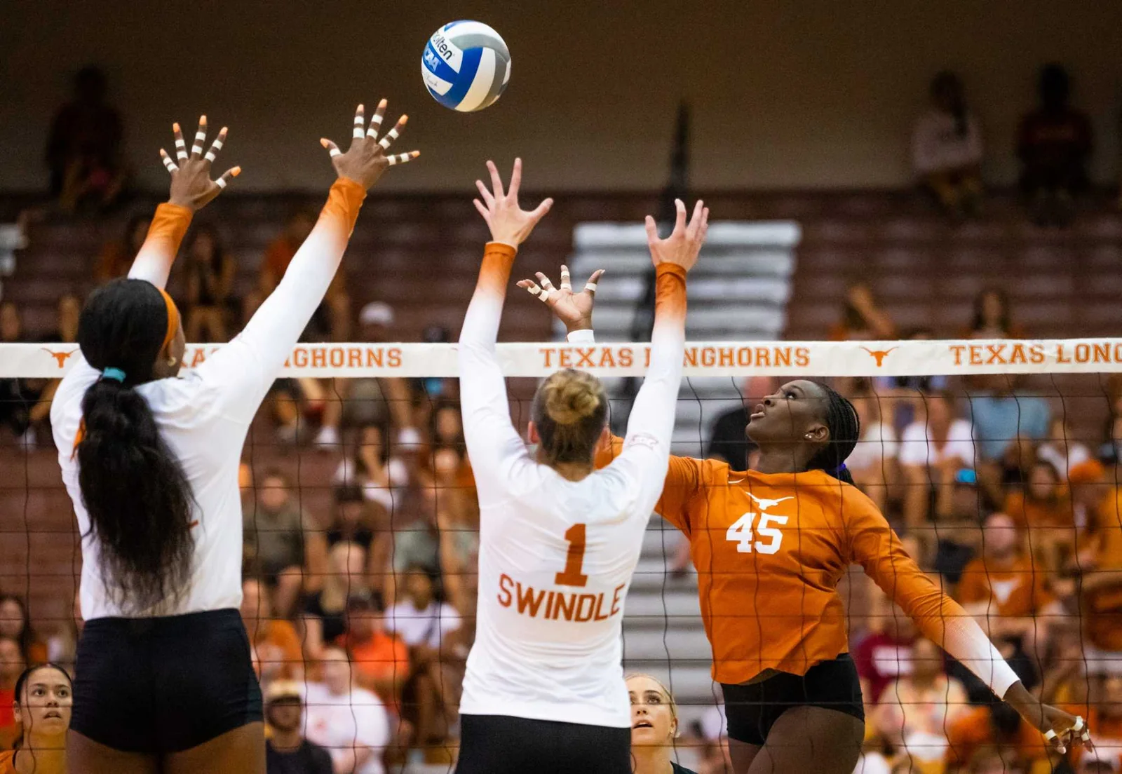 Texas volleyball hitter in orange attacking against two white-jersey blockers