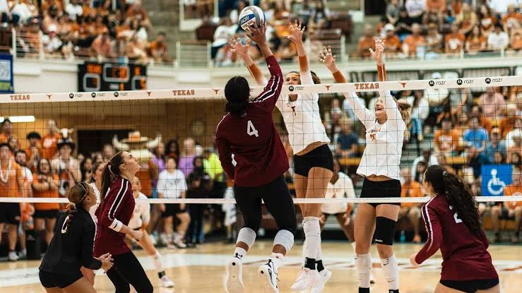 Two Texas volleyball players in white jerseys blocking an attack by a maroon-clad opponent
